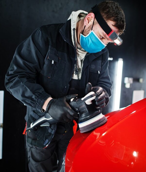 Car detailing concept. Man in face mask with orbital polisher in repair shop polishing orange suv car.