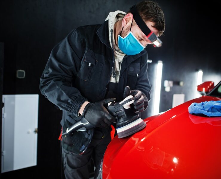 Car detailing concept. Man in face mask with orbital polisher in repair shop polishing orange suv car.